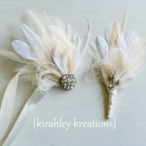 May include: Two white feather bouquets with crystal embellishments. The larger bouquet has a larger crystal and is attached to a white satin ribbon. The smaller bouquet has three smaller crystals.