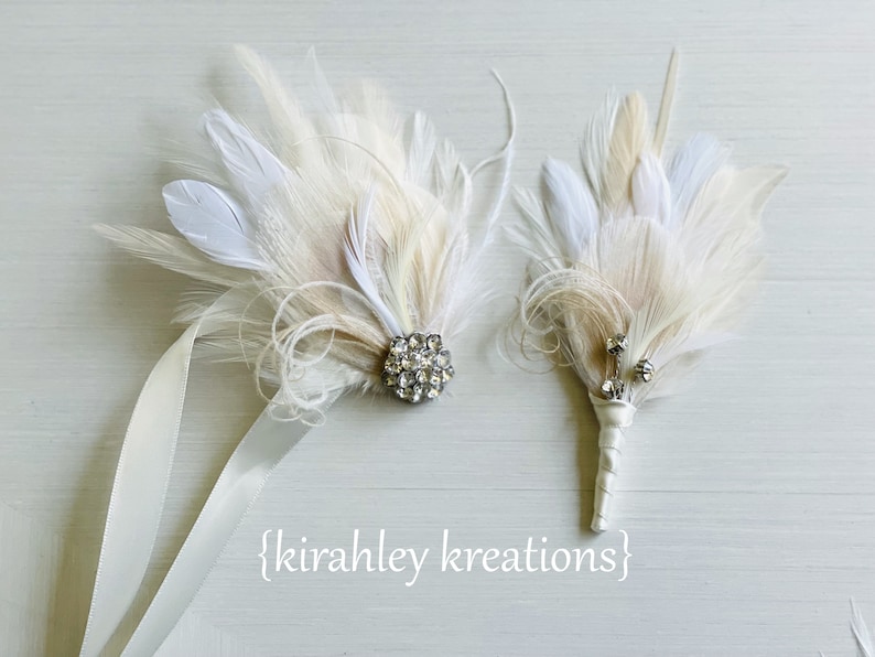 May include: Two white feather bouquets with crystal embellishments. The larger bouquet has a larger crystal and is attached to a white satin ribbon. The smaller bouquet has three smaller crystals.