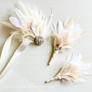 Puede incluir: Tres boutonni&egrave;res de plumas marfil con adornos de cristal. Las boutonni&egrave;res est&aacute;n atadas con una cinta de sat&eacute;n marfil.