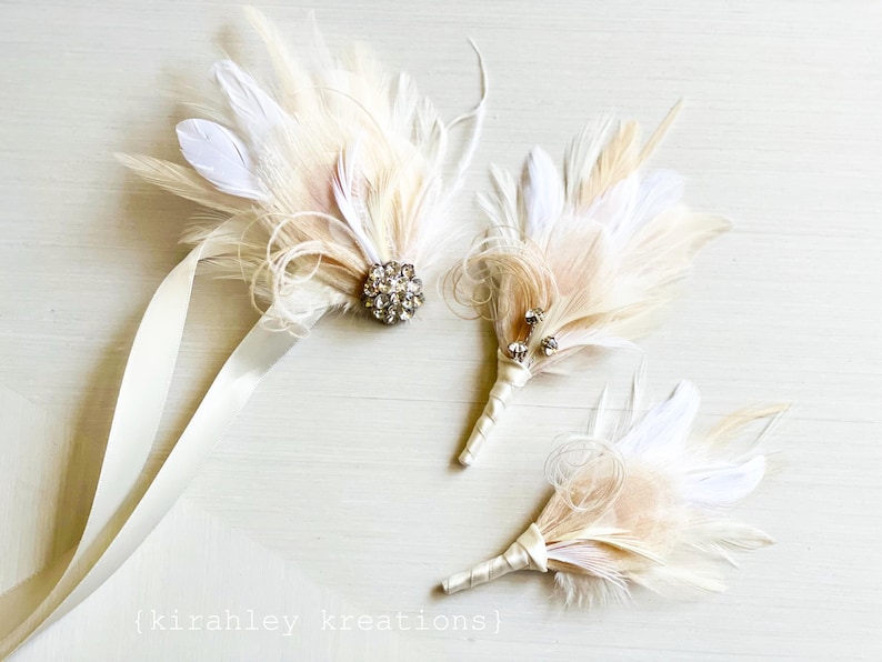 Puede incluir: Tres boutonni&egrave;res de plumas marfil con adornos de cristal. Las boutonni&egrave;res est&aacute;n atadas con una cinta de sat&eacute;n marfil.