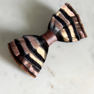 May include: A handcrafted bow tie featuring a feather design with brown, black, and gold tones. The bow tie has a brown band in the center. The text "kirahley kreations" is at the bottom.