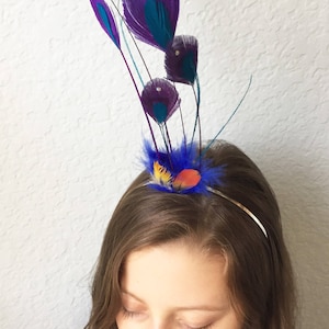May include: A decorative headband featuring purple and teal peacock feathers. The feathers are accented with small gold beads and arranged above a cluster of blue, yellow, and orange feathers. The text "{kirahley kreations}" is visible in the upper right corner.