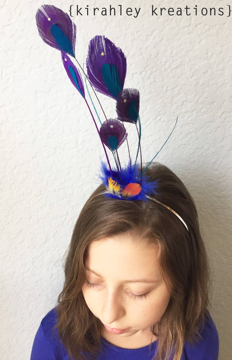 May include: A decorative headband featuring purple and teal peacock feathers. The feathers are accented with small gold beads and arranged above a cluster of blue, yellow, and orange feathers. The text "{kirahley kreations}" is visible in the upper right corner.