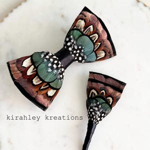 May include: A handcrafted feather bow tie and boutonniere set. The bow tie features brown, green, and white feathers with a black satin band. The boutonniere has a similar design. The set is displayed on a white marble surface.