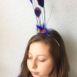 May include: A decorative headband featuring purple and teal peacock feathers with rhinestone accents. The headband has a blue, orange, and yellow feather accent. The text "{kirahley kreations}" is visible at the bottom of the image.