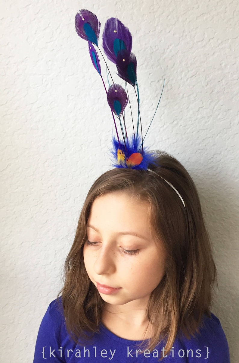 May include: A decorative headband featuring purple and teal peacock feathers with rhinestone accents. The headband has a blue, orange, and yellow feather accent. The text "{kirahley kreations}" is visible at the bottom of the image.