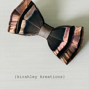 May include: A handcrafted bow tie made of feathers in shades of brown, black, and copper. The bow tie is held together by a brown leather band. The text "brown leather" and "{kirahley kreations}" are visible in the image.