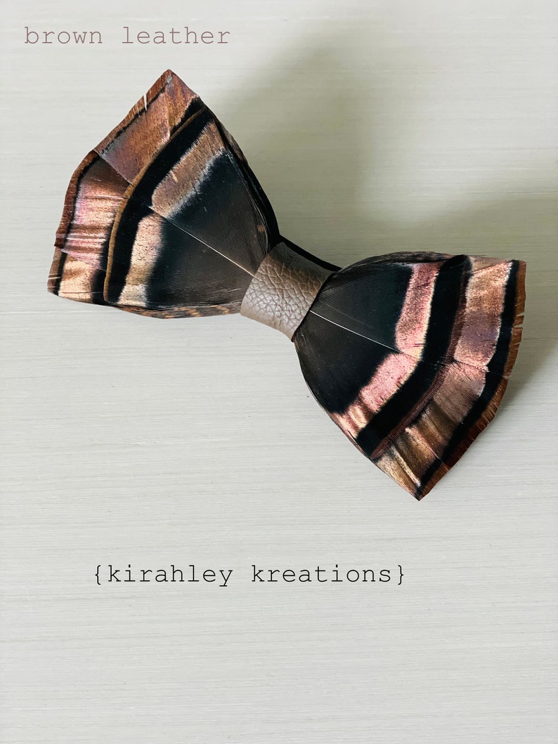 May include: A handcrafted bow tie made of feathers in shades of brown, black, and copper. The bow tie is held together by a brown leather band. The text "brown leather" and "{kirahley kreations}" are visible in the image.