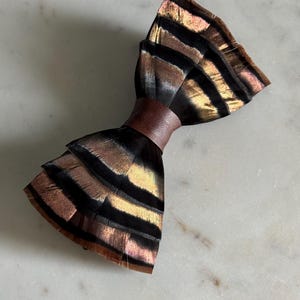 May include: A handmade bow tie made of iridescent feathers in shades of black, brown, and gold, with a brown leather band. The bow tie is displayed on a white marble surface. The text "kirahley kreations" is at the bottom.