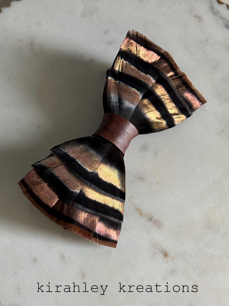 May include: A handmade bow tie made of iridescent feathers in shades of black, brown, and gold, with a brown leather band. The bow tie is displayed on a white marble surface. The text "kirahley kreations" is at the bottom.