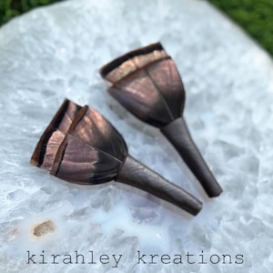 May include: Two dark brown, tulip-shaped earrings with a metallic sheen, resting on a white, marbled surface. The earrings have a textured appearance and are displayed against a blurred green background. The text "kirahley kreations" is visible.