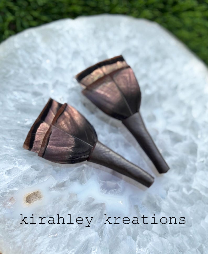 May include: Two dark brown, tulip-shaped earrings with a metallic sheen, resting on a white, marbled surface. The earrings have a textured appearance and are displayed against a blurred green background. The text "kirahley kreations" is visible.