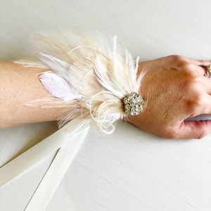 Puede incluir: Un brazalete de mu&ntilde;eca de plumas blancas con un adorno de cristal y una cinta de sat&eacute;n blanca.
