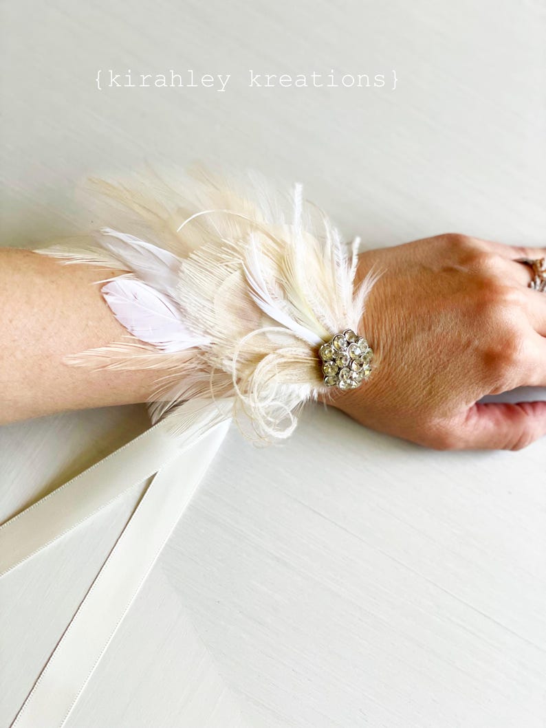 Puede incluir: Un brazalete de mu&ntilde;eca de plumas blancas con un adorno de cristal y una cinta de sat&eacute;n blanca.
