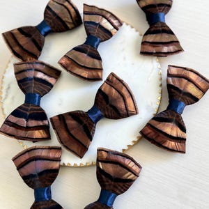 May include: A collection of handmade bow ties with a feather design. The bow ties feature brown and black feathers with a blue satin ribbon. The bow ties are arranged on a white plate with a gold trim. The text "kirahley kreations" is at the top.