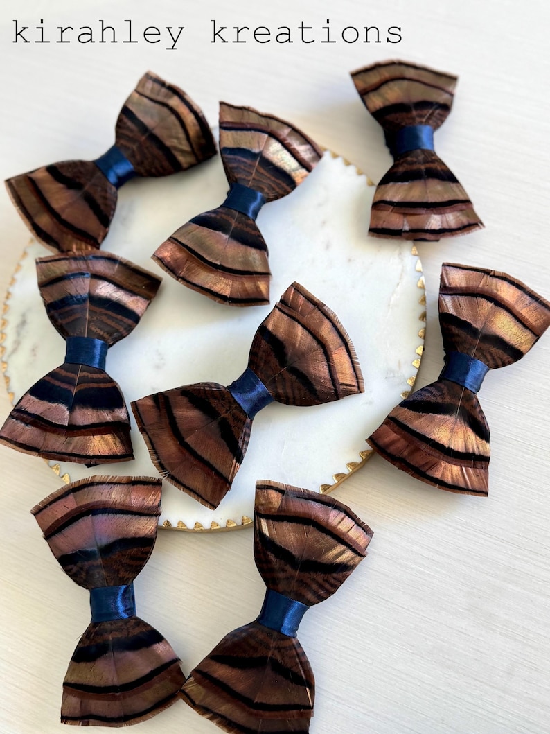 May include: A collection of handmade bow ties with a feather design. The bow ties feature brown and black feathers with a blue satin ribbon. The bow ties are arranged on a white plate with a gold trim. The text "kirahley kreations" is at the top.
