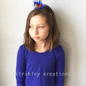 May include: A young person wearing a blue long-sleeved top and a decorative headband with purple and teal feathers. The text "{kirahley kreations}" is visible on the shirt.