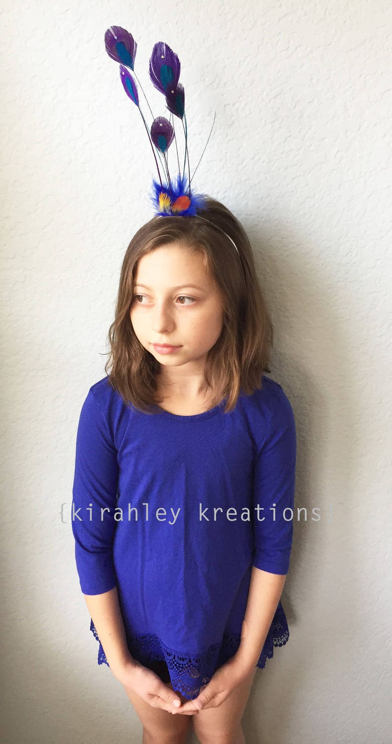 May include: A young person wearing a blue long-sleeved top and a decorative headband with purple and teal feathers. The text "{kirahley kreations}" is visible on the shirt.