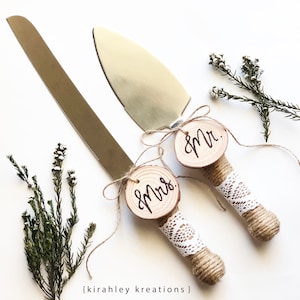 May include: A set of two cake cutting knives with wooden handles wrapped in twine and lace. The handles are decorated with round wood slices that say "Mr." and "Mrs." in calligraphy.