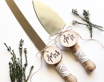 May include: A set of two cake cutting knives with wooden handles wrapped in twine and lace. The handles are decorated with round wood slices that say "Mr." and "Mrs." in calligraphy.