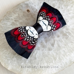 Puede incluir: Pajarita de plumas hecha a mano con una banda de satén negro. La pajarita presenta plumas rojas, blancas y negras con un patrón moteado. El texto "kirahley kreations" es visible en la superficie.