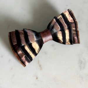 May include: A handcrafted bow tie made from feathers, featuring a brown leather band. The feathers display a pattern of black and gold stripes. The text "kirahley kreations" is visible at the bottom.