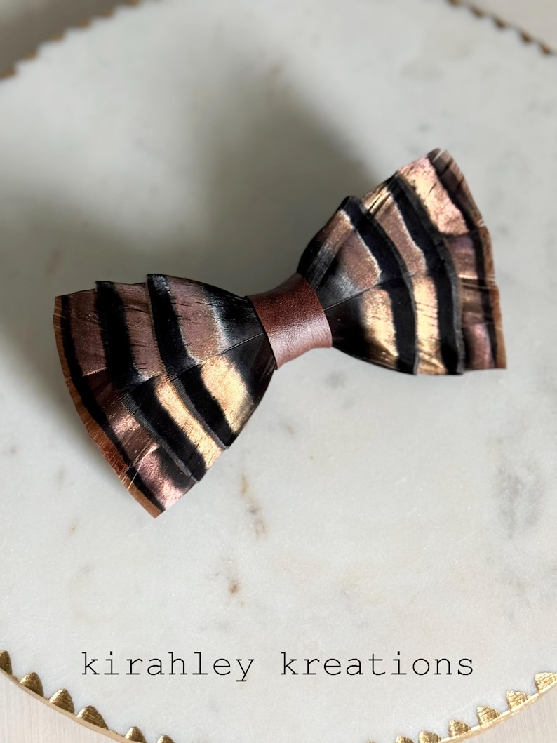 May include: A handcrafted bow tie made from feathers, featuring a brown leather band. The feathers display a pattern of black and gold stripes. The text "kirahley kreations" is visible at the bottom.