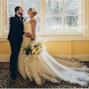 May include: A bride and groom stand in front of a window, smiling at each other. The bride is wearing a white lace wedding dress with a long train and a feather headpiece. The groom is wearing a black suit with a white bow tie. The couple is holding a bouquet of white flowers.