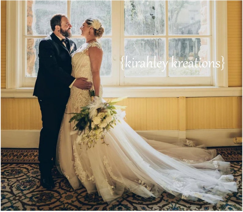 May include: A bride and groom stand in front of a window, smiling at each other. The bride is wearing a white lace wedding dress with a long train and a feather headpiece. The groom is wearing a black suit with a white bow tie. The couple is holding a bouquet of white flowers.
