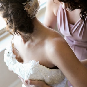 May include: A bride is getting ready for her wedding day, with a bridesmaid helping her adjust her white lace wedding dress. The bride is wearing a delicate feather hairpiece.