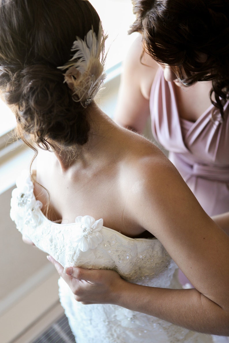 May include: A bride is getting ready for her wedding day, with a bridesmaid helping her adjust her white lace wedding dress. The bride is wearing a delicate feather hairpiece.