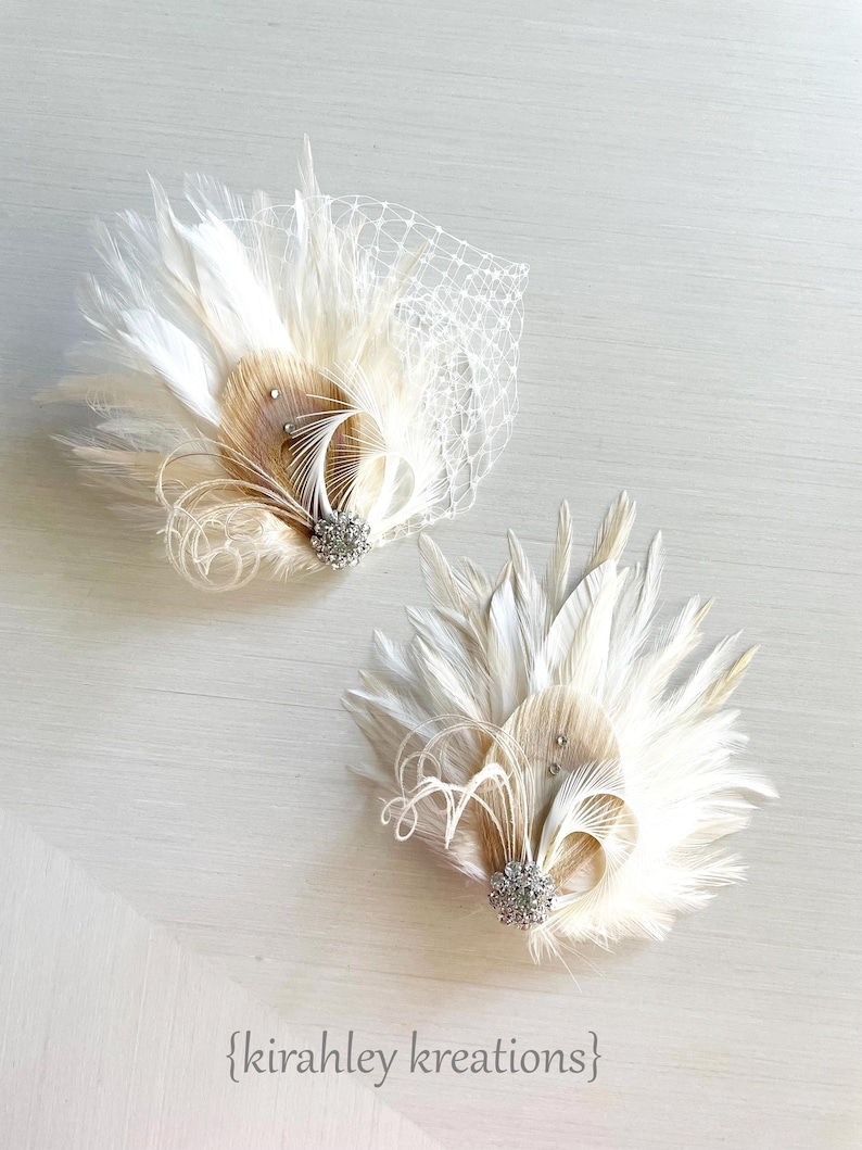 Op de afbeelding: Twee decoratieve haarspeldjes met witte en beige veren, zilverkleurige strass-accenten en een witte netsluier op &eacute;&eacute;n stuk. De tekst {kirahley kreations} staat onderaan de afbeelding gedrukt in een eenvoudig zwart lettertype.
