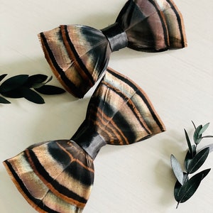 Puede incluir: Dos pajaritas de plumas marrones y negras con detalles de cinta de satén negra. Las pajaritas están dispuestas sobre una superficie blanca con hojas verdes.