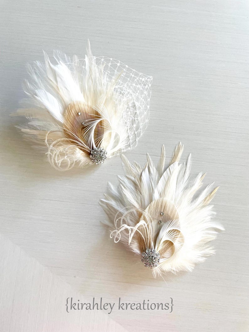 May include: Two ivory feather hair clips with a rhinestone embellishment. Each clip features a fan of feathers with a smaller cluster of feathers in the center.