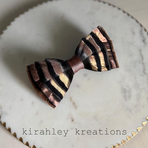 May include: A handcrafted bow tie with a unique feather design. The bow tie features a brown satin center and is adorned with feathers in shades of brown, black, and gold. The text "kirahley kreations" is visible on the surface.