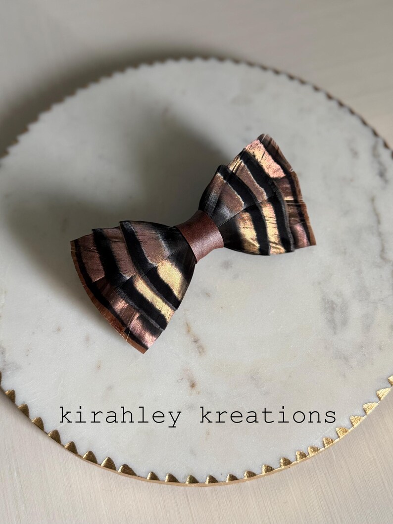 May include: A handcrafted bow tie with a unique feather design. The bow tie features a brown satin center and is adorned with feathers in shades of brown, black, and gold. The text "kirahley kreations" is visible on the surface.