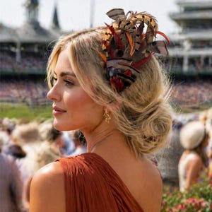 May include: A woman with blonde hair styled in a low bun, wearing a rust-colored dress and a feathered headpiece. The headpiece features brown, orange, and red feathers. The background shows a racetrack and spectators.