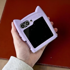 Puede incluir: Una funda de teléfono lavanda con orejas de gato, sostenida en una mano. La funda tiene una pantalla rectangular y dos lentes de cámara. La funda del teléfono es de color lavanda liso.