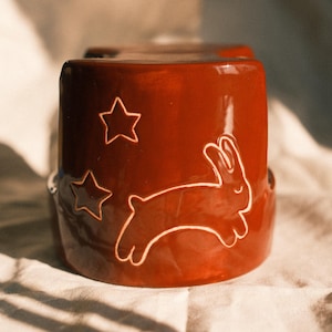 Può includere: Una tazza cilindrica in ceramica color arancione scuro lucido con il contorno chiaro disegnato a mano di un coniglio che salta e due stelle a cinque punte sul lato. La tazza presenta una superficie liscia e riflettente.