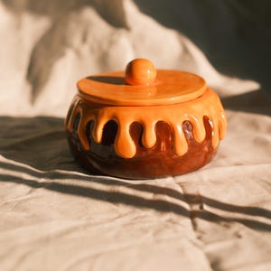Puede incluir: Un tarro de cerámica redondo con una base de color marrón oscuro y una tapa naranja brillante. La tapa tiene un pomo central y la parte superior del tarro está decorada con un esmalte naranja texturizado que gotea como un líquido.