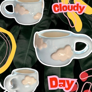 Pode incluir: Três canecas de cerâmica azul claro com desenhos de nuvens brancas em relevo. As canecas estão dispostas sobre um fundo de folhas verdes escuras com formas abstratas coloridas e as palavras "cloudy" e "day" em letras vermelhas e rosa em negrito.