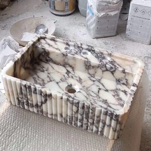 May include: A white marble sink with a rippled edge and a grey veining pattern. The sink has two small holes and a larger hole in the centre.