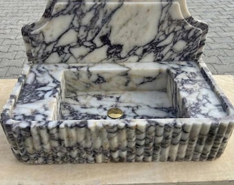 May include: A rectangular marble sink with a decorative back and a fluted front. The sink features a white and gray veined pattern, a rectangular basin, and a gold-colored drain. The sink is set on a wooden surface.
