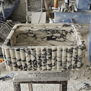 May include: A rectangular marble sink with a white and black veined pattern. The sink has a textured, ribbed exterior. The image is taken in a workshop setting, surrounded by tools and construction materials.