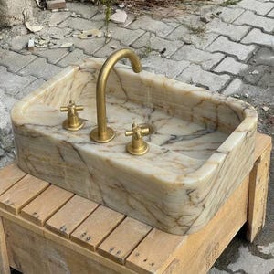 May include: Rectangular marble sink with a gold-coloured tap and two matching handles. The sink has a beige and brown veined pattern. The tap is curved, and the handles are cross-shaped.
