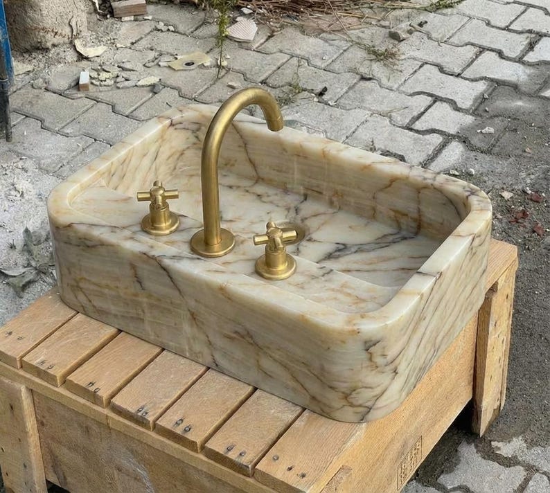 May include: Rectangular marble sink with a gold-coloured tap and two matching handles. The sink has a beige and brown veined pattern. The tap is curved, and the handles are cross-shaped.