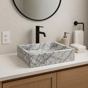 May include: White marble vessel sink with a black faucet and a white countertop. The sink is rectangular and has a square shape. The faucet is tall and has a single handle.