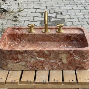 May include: A rectangular red marble sink with gold-coloured tap and handles. The sink is set on a wooden pallet. The marble has a natural pattern of white and red veins.