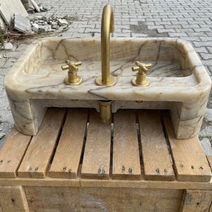 May include: A rectangular marble sink with a brass tap and two brass handles. The sink is set on a wooden base. The marble has a beige and brown veined pattern.