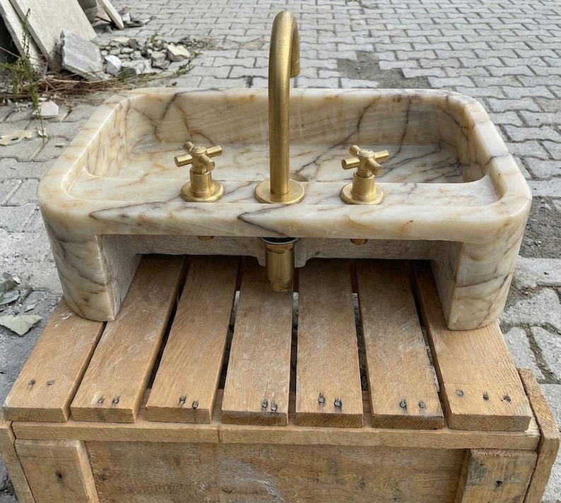 May include: A rectangular marble sink with a brass tap and two brass handles. The sink is set on a wooden base. The marble has a beige and brown veined pattern.
